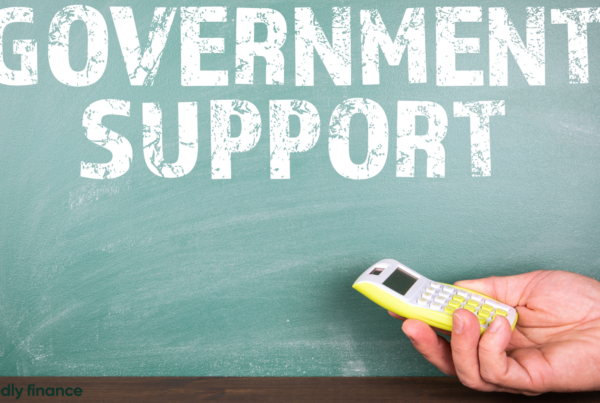 a chalk board with the words "Government Support" written; a visible hand holding a calculator is rested below